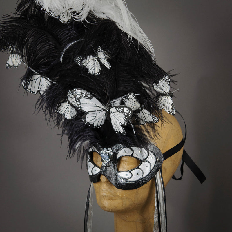 Beautiful Masquerade Mask, crested in Black and White Ostrich Plumes and assorted plumage. White feather monarch butterflies flutter on the crest. Embellished with Swarovski crystals, dichroic glass and polished black quartz.  Hand made in the USA using traditional Venetian paper-mache technique. Lined with hypoallergenic stretch velvet for comfort. Side view.