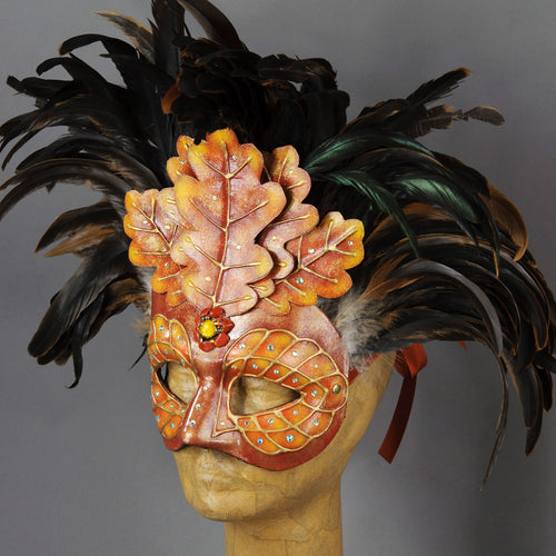 Beautiful Masquerade Mask, crested in Black coque feathers accented with red and gold handmade oak leaves. Embellished with Swarovski crystals, dichroic glass and polished stones. This is one of our versions of the Green Man mask in shades of red and gold to reflect the autumn/fall.  Hand made in the USA using traditional Venetian paper-mache technique. Lined with hypoallergenic stretch velvet for comfort. Side view.