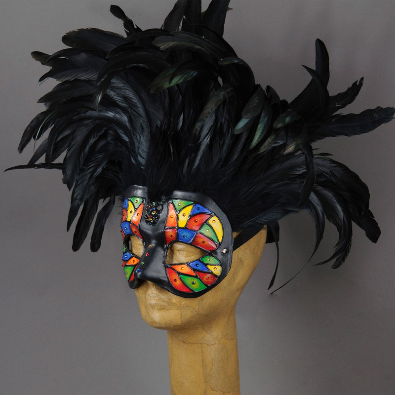 Beautiful Masquerade Mask, crested in Black coque feathers. Stained glass motif painted in rainbow colors and embellished with Swarovski crystals, dichroic glass and polished stones. Shades of red, orange, yellow, green and blue.  Hand made in the USA using traditional Venetian paper-mache technique. Lined with hypoallergenic stretch velvet for comfort. Side view.